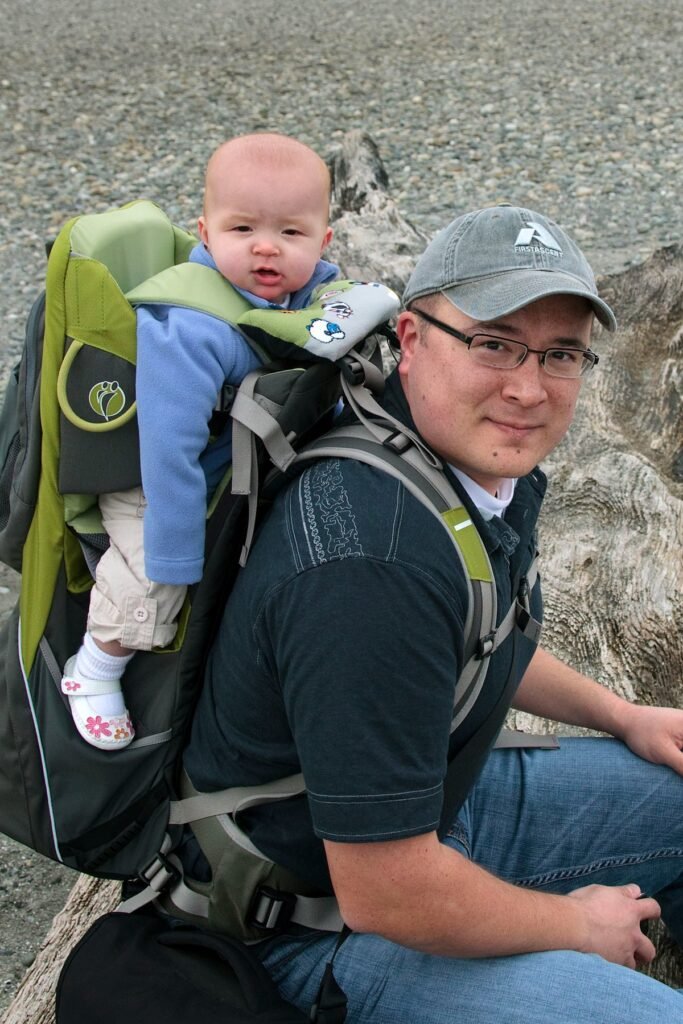A parent carries a baby in a green and gray hiking backpack while exploring nature.