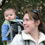 A woman carrying a toddler in a front carrier during a spring hike, with blooming trees in the background.