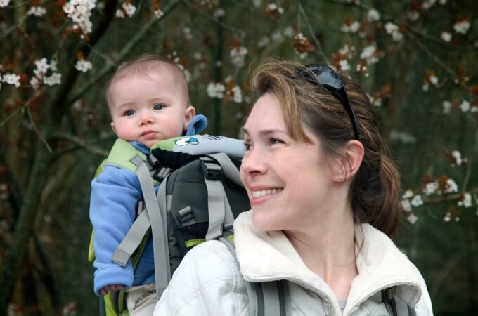 A woman carrying a toddler in a front carrier during a spring hike, with blooming trees in the background.