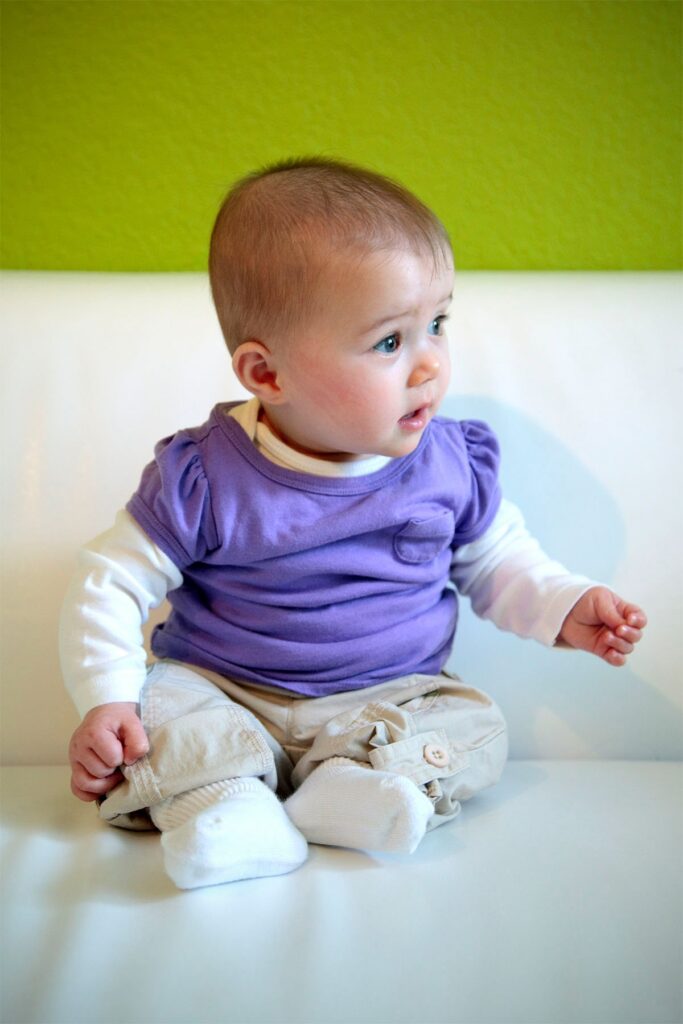 A baby wearing a purple and white outfit sits on a white surface. Bright green wall provides a vibrant backdrop.