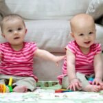 Twin babies in pink striped shirts sitting on the floor playing with colorful toys near a couch.