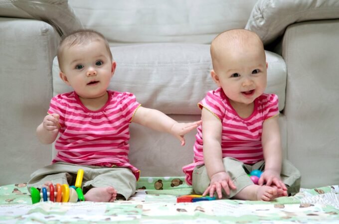 Twin babies in pink striped shirts sitting on the floor playing with colorful toys near a couch.