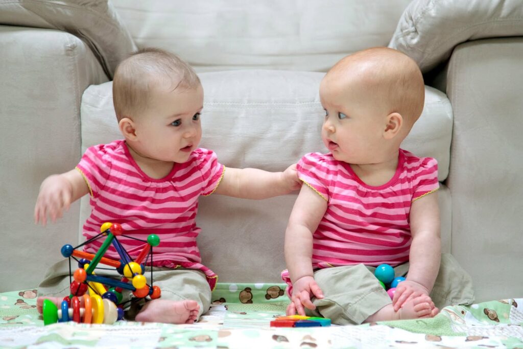 Two babies in pink striped shirts sit on a play mat, playing with magnetic blocks and colorful toys in front of a couch.