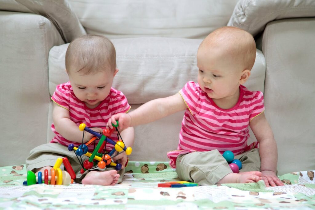 Two babies in matching pink striped shirts playing with a colorful building toy on the floor.