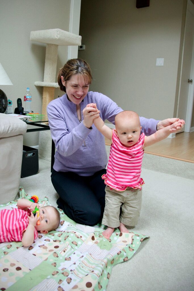 A woman supports a toddler as they take their first steps on a patterned blanket.