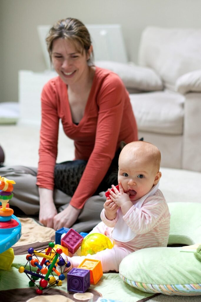A smiling adult in a red top plays on the floor with a baby surrounded by colorful building blocks and toys, with a beige sofa in the background.