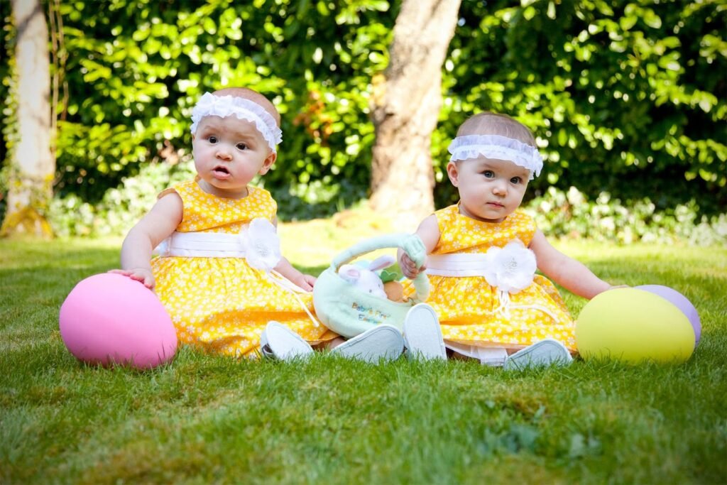 Two young children in yellow outfits sit on a sunny lawn with colorful balloons.