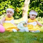 Two young children in yellow outfits sit on a sunny lawn with colorful balloons.