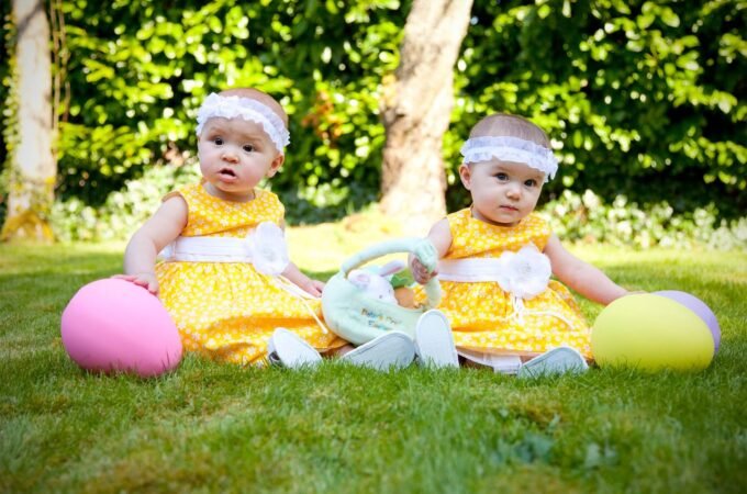 Two young children in yellow outfits sit on a sunny lawn with colorful balloons.