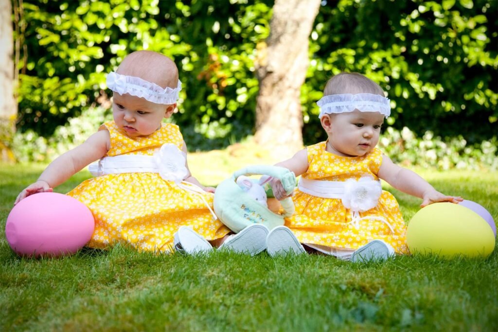 Two babies dressed in yellow dresses sit outdoors on the grass, each holding a large pink or yellow decorative egg.