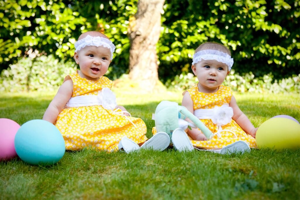 Two babies in yellow dresses playing with pastel Easter eggs on a sunny lawn.