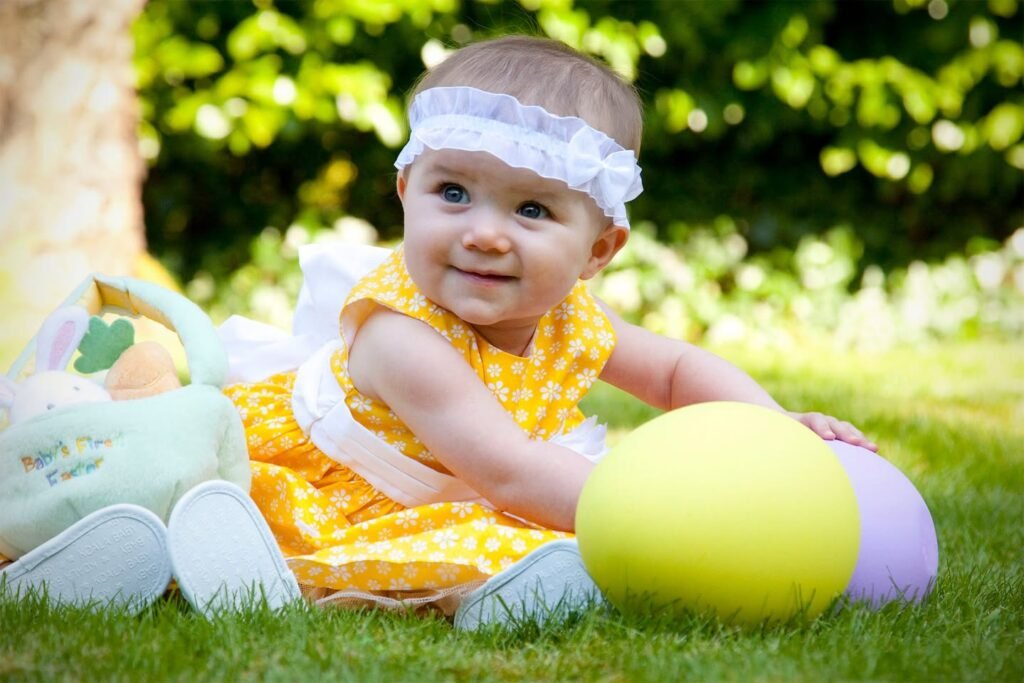 A baby sitting in the grass during an Easter egg hunt. The baby is wearing a yellow dress and a white headband.