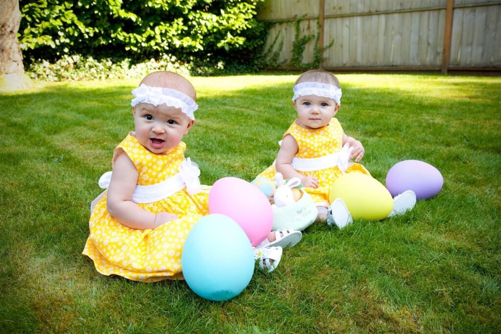 Two toddlers in yellow outfits sit on grass holding pastel Easter eggs.