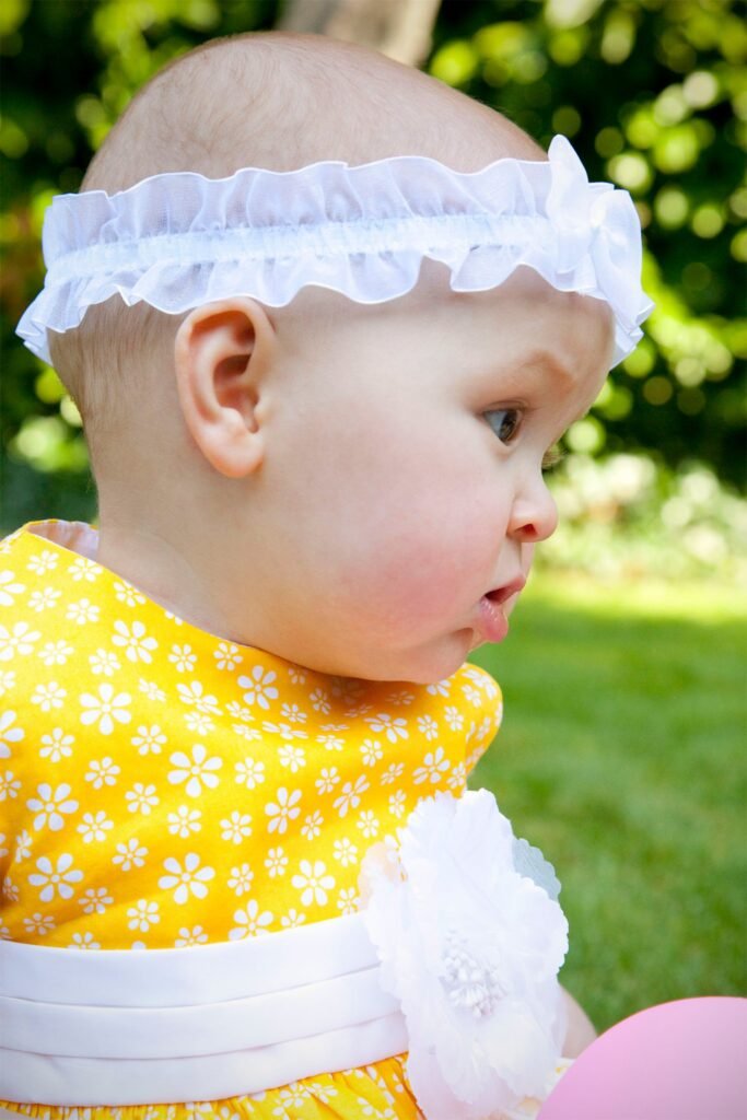 A baby wearing a yellow floral dress and a white ruffled headband outdoors.
