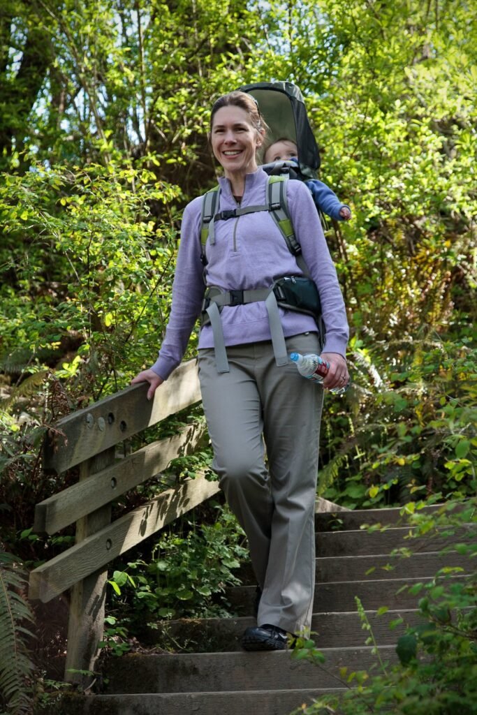 A smiling person walks down a wooded trail carrying a baby in a back carrier, surrounded by dense green forest.