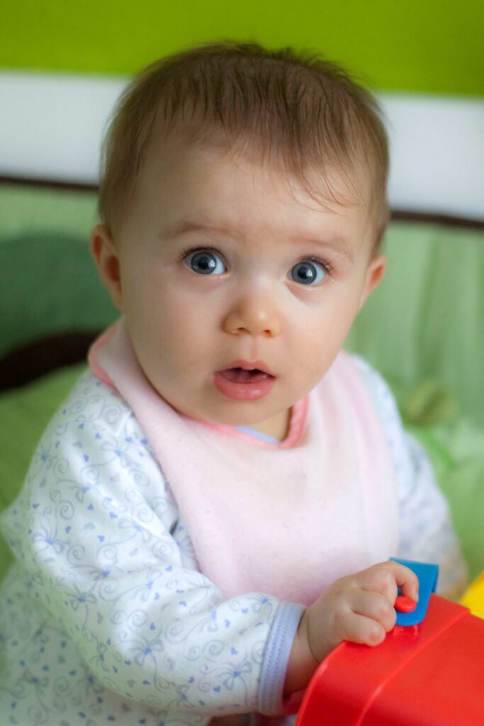 A close-up of a baby in a high chair holding a red and blue toy tool. The baby wears a pink bib and looked surprised while playing.