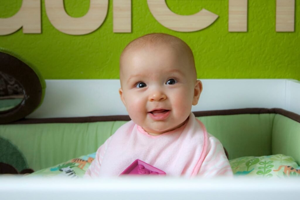 A baby smiling inside a crib with light green walls and wooden letters mounted above.
