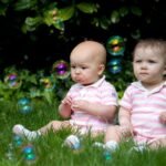 Two small children sitting on green grass surrounded by floating soap bubbles.