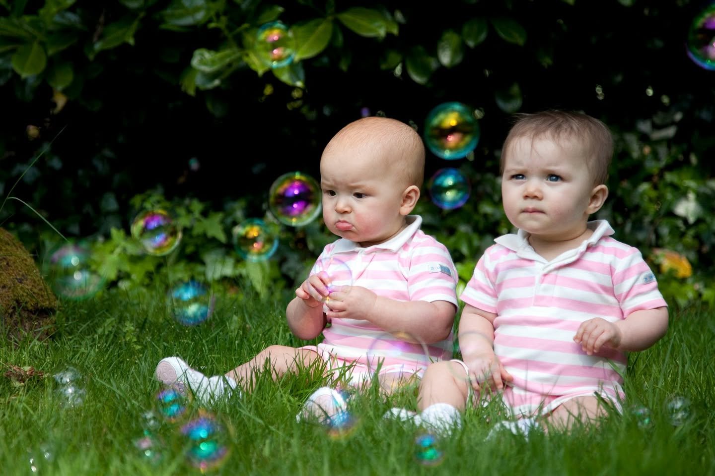 May Days Two small children sitting on green grass surrounded by floating soap bubbles.