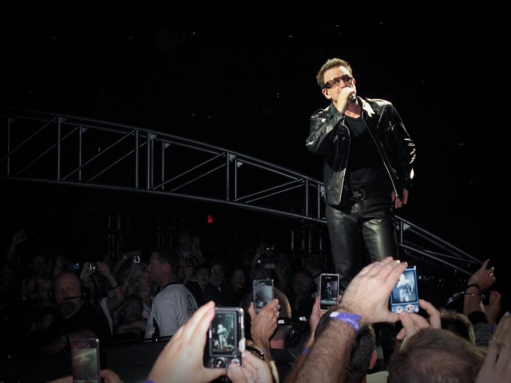 A performer on stage in a black leather jacket and sunglasses sings to a crowd while many audience members record the moment with mobile phones.