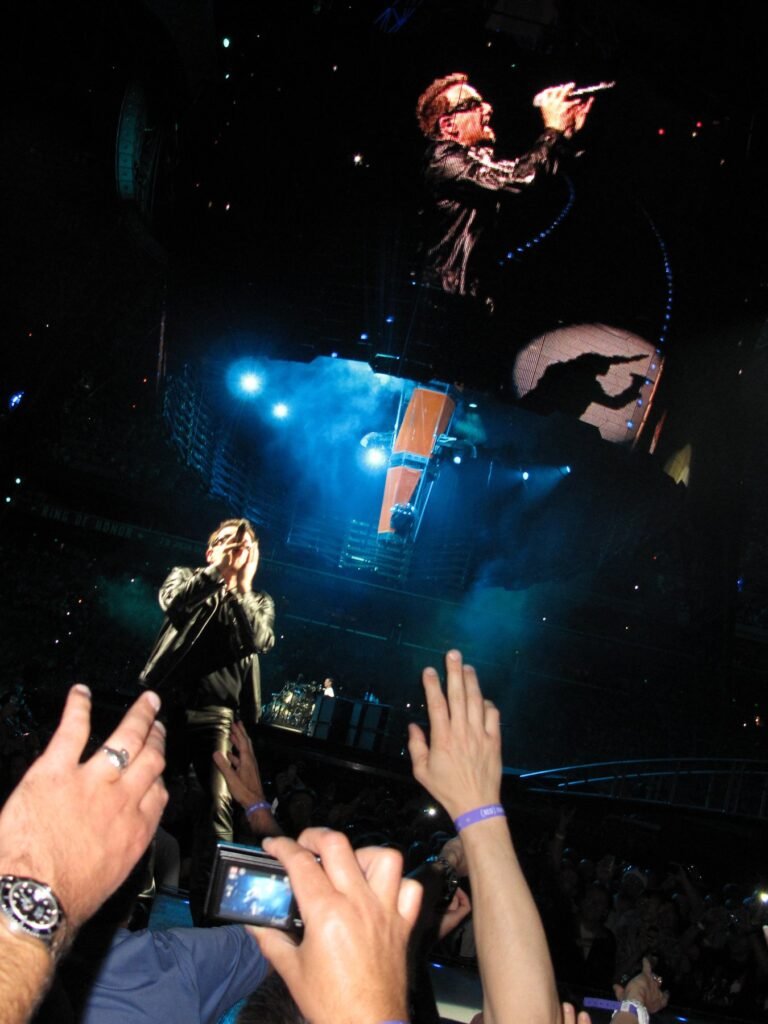 Performers on stage under blue lighting with fans holding up phones and cameras.