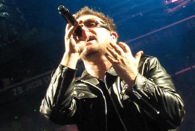 A performer in a black leather jacket and sunglasses sings into a microphone under blue stage lighting at a live concert.