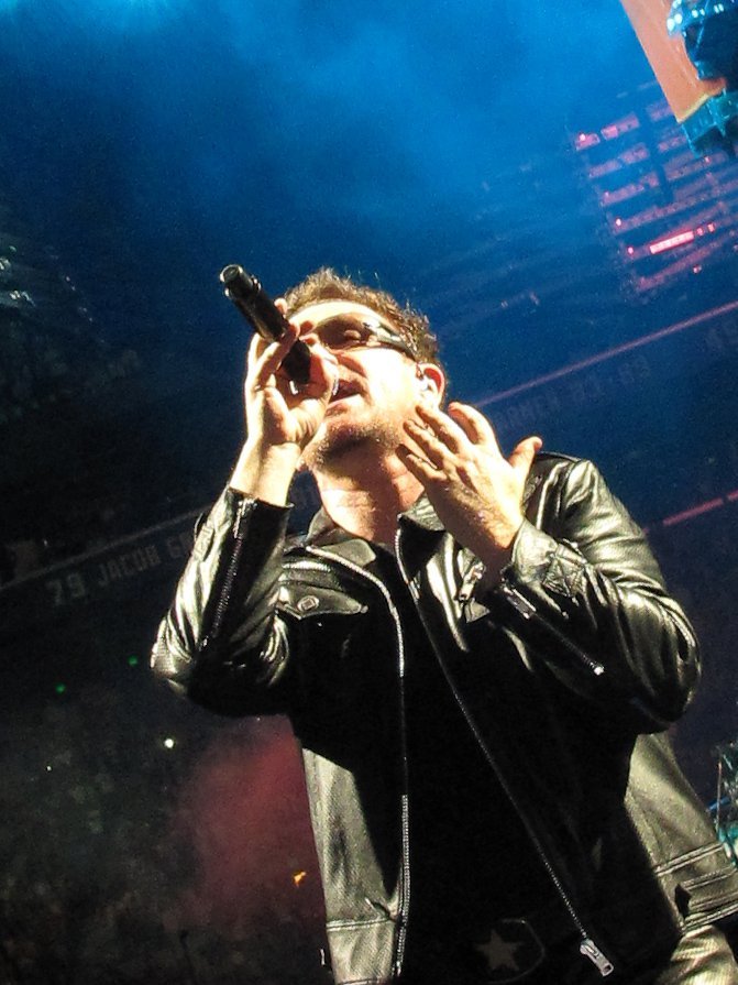 A performer in a black leather jacket and sunglasses sings into a microphone under blue stage lighting at a live concert.