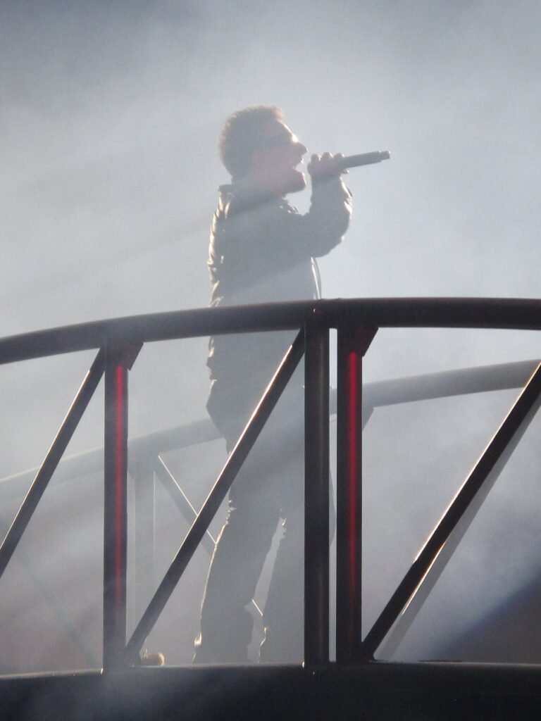 A performer sings into a microphone on a stage surrounded by fog, creating a dramatic silhouette against the metallic railing.