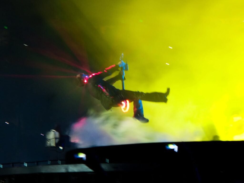 A performer mid-air on stage illuminated by bright yellow and red lights. The dynamic scene captures a live concert show.