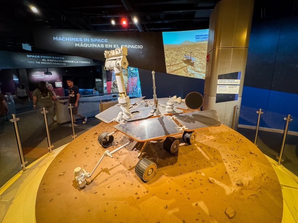 A detailed model of a Mars rover displayed on a rocky surface, set within an exhibit labeled 'Machines in Space.'