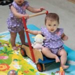 Ross Lake Backpacking Two toddlers in matching lavender outfits play with a wooden wagon toy on a colorful rug.