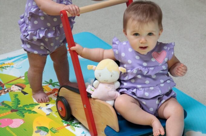 Two toddlers in matching lavender outfits play with a wooden wagon toy on a colorful rug.