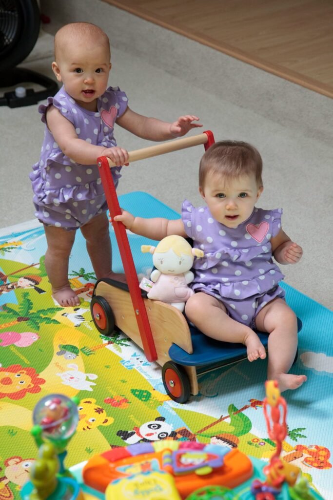 Two toddlers in matching lavender outfits play with a wooden wagon toy on a colorful rug.