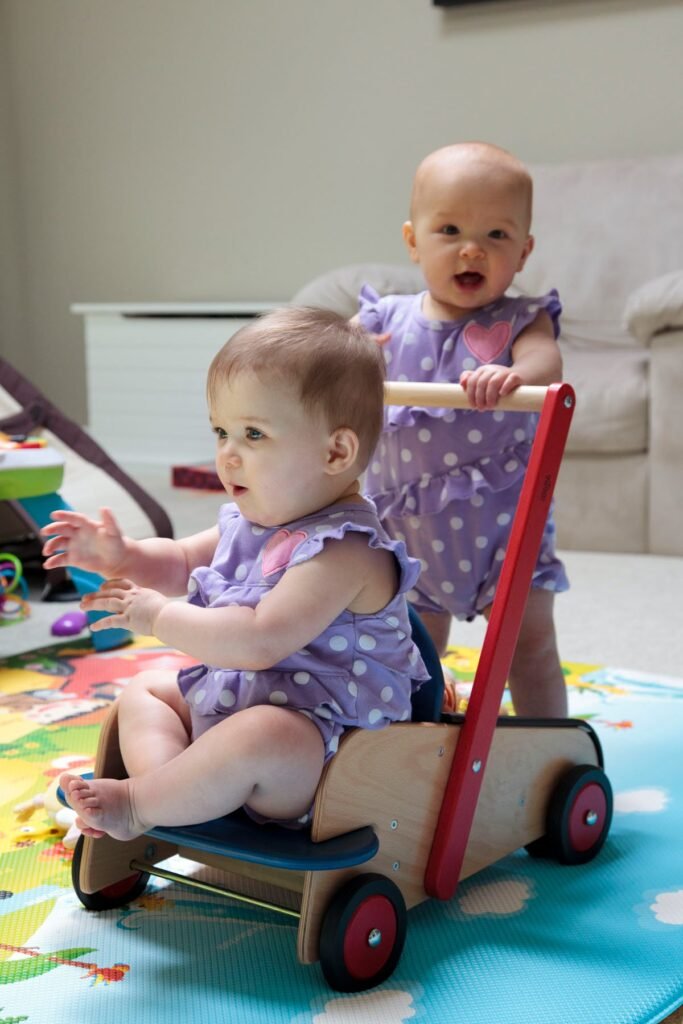 A baby rides a wooden pull-along wagon while another infant pushes from behind on a colorful play mat.