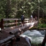 Hikers with backpacks cross a wooden footbridge over a rushing river in a dense forest.