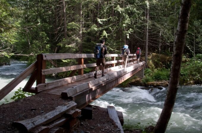Hikers with backpacks cross a wooden footbridge over a rushing river in a dense forest.