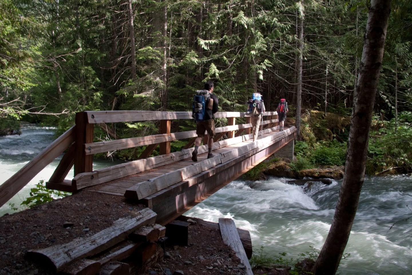 Ross Lake Backpacking Hikers with backpacks cross a wooden footbridge over a rushing river in a dense forest.