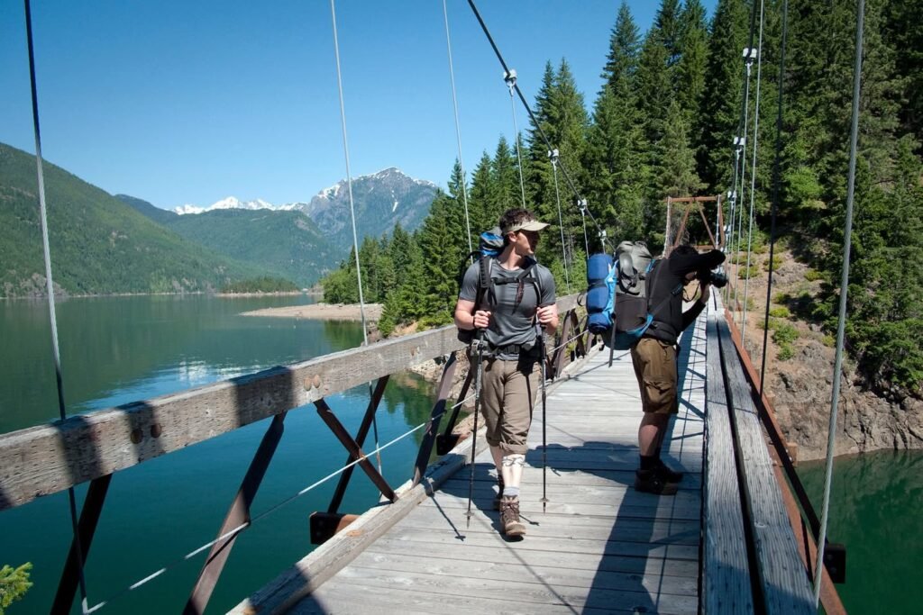 Hikers crossing a wooden suspension footbridge over a turquoise lake with evergreen trees and mountains in the distance under a clear blue sky.