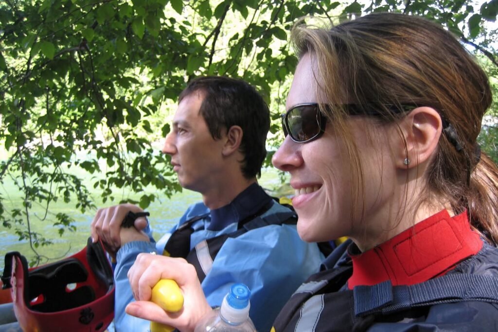 Two travelers pause on an outdoor excursion near a body of water to enjoy a fresh lemon snack. Their gear includes backpacks and waterproof clothing, set against green foliage.