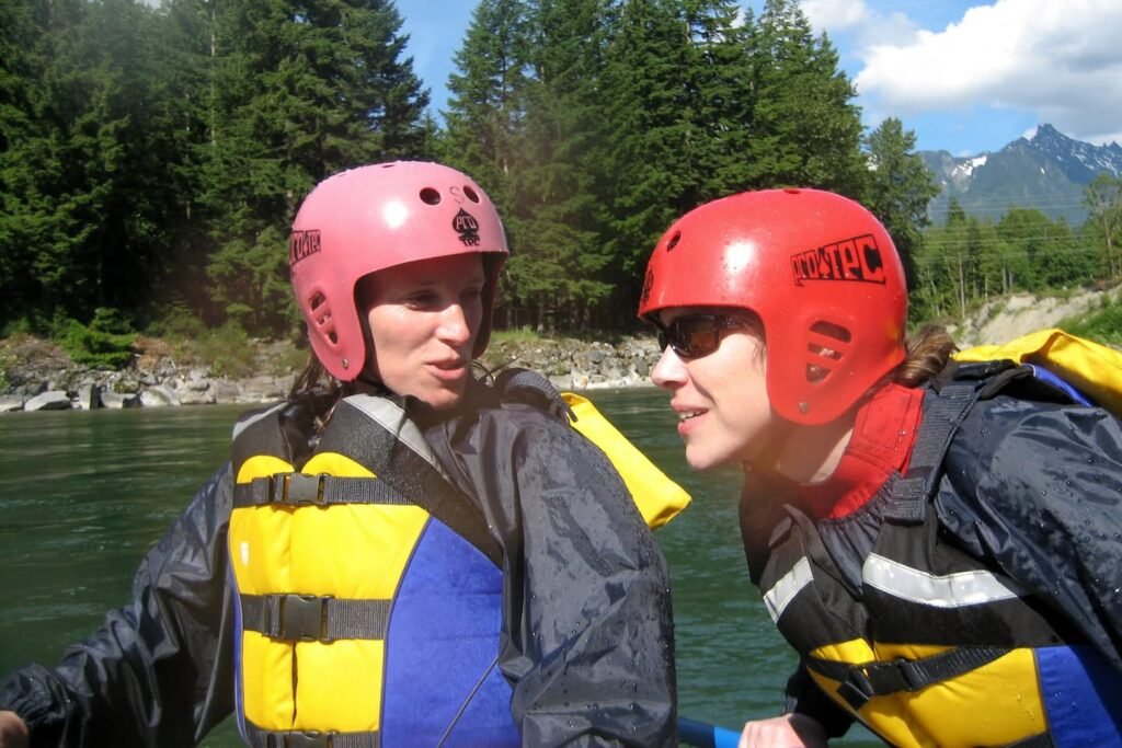 Two people wearing helmets and life jackets enjoy a rafting trip on a river surrounded by mountains and trees. The vibrant gear contrasts with the natural scenery.