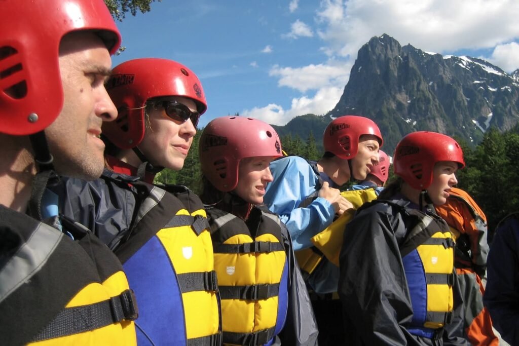 A group in red helmets and life jackets prepares for a river adventure against a scenic mountain backdrop.
