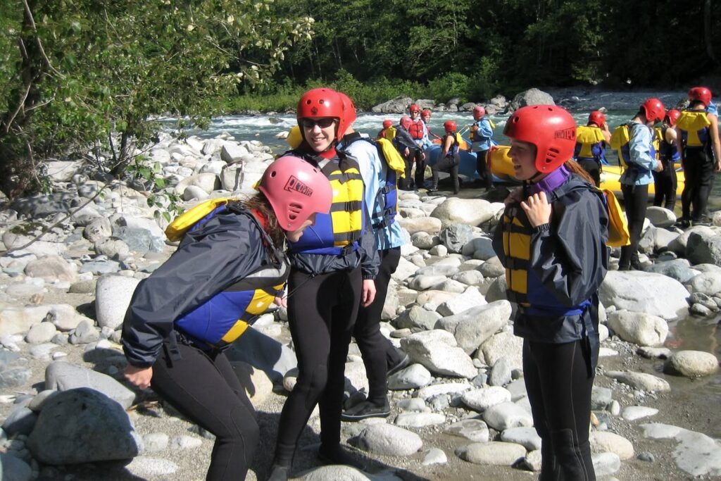 A group of children wearing helmets and life jackets gather on a rocky riverbank, preparing for a water-based activity.