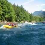 A group in a yellow raft navigating rushing river rapids. Lush evergreen trees line the riverbanks under a bright sky.