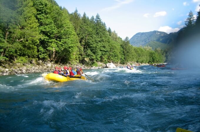 A group in a yellow raft navigating rushing river rapids. Lush evergreen trees line the riverbanks under a bright sky.