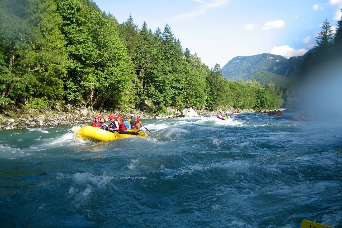Skykomish River White Water Rafting A group in a yellow raft navigating rushing river rapids. Lush evergreen trees line the riverbanks under a bright sky.
