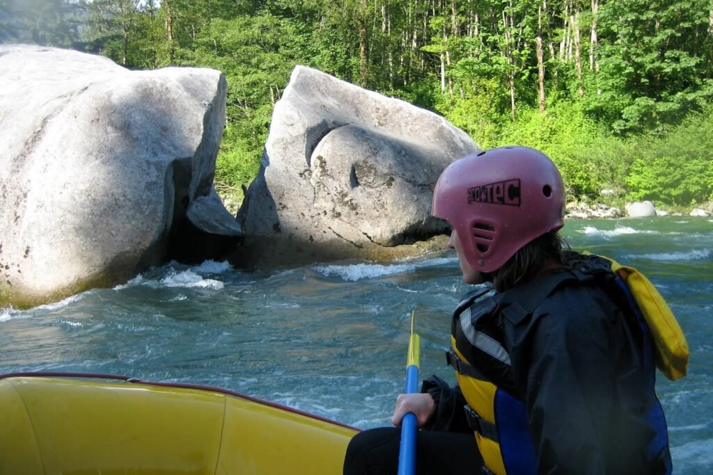 Paddling a yellow raft through a river with large rocks and green forest in the background.