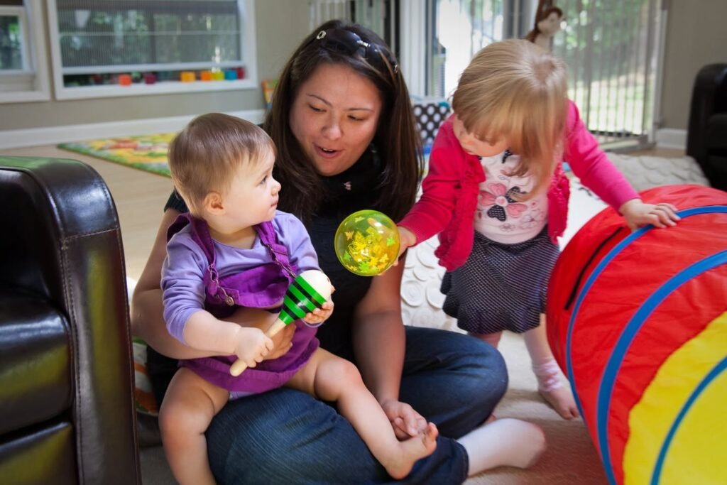 A caregiver holds a baby while a toddler plays with colorful indoor gym equipment.