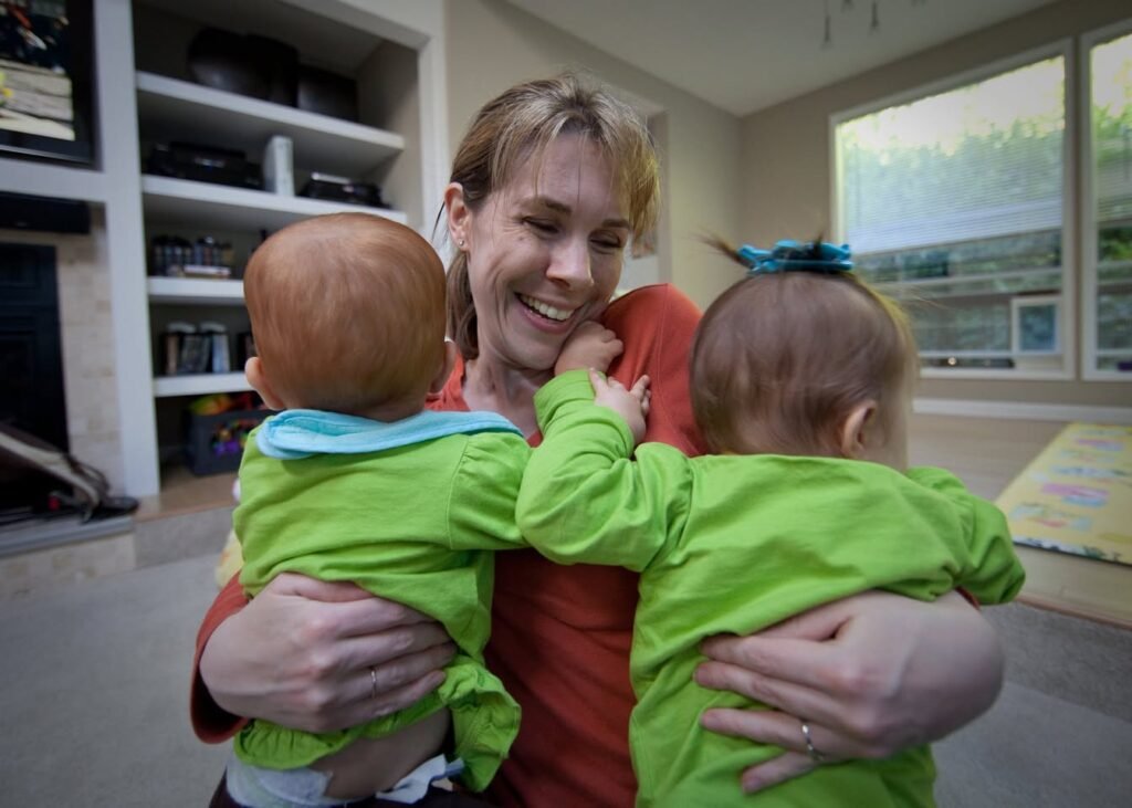A smiling person holds two babies in matching green outfits inside a bright living room with large windows.