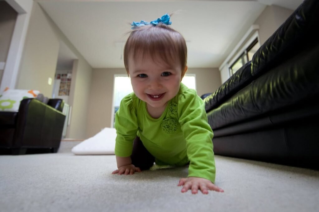 A happy toddler crawls across the carpet in a bright living room, wearing a green shirt.