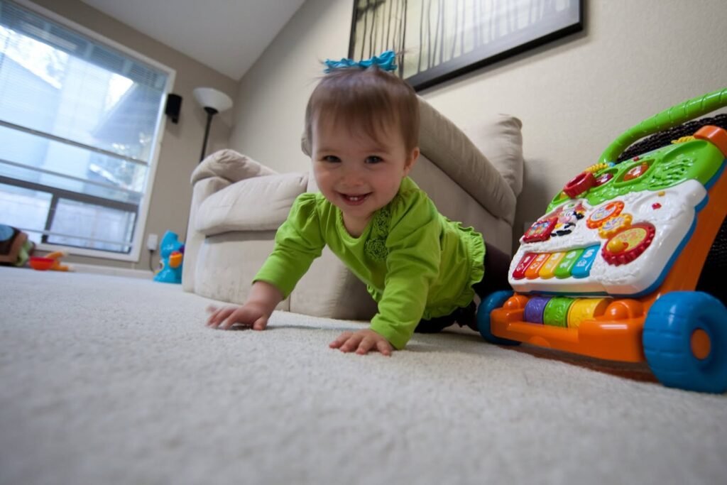 A young child in a green shirt smiles while crawling on a carpet near a toy car.